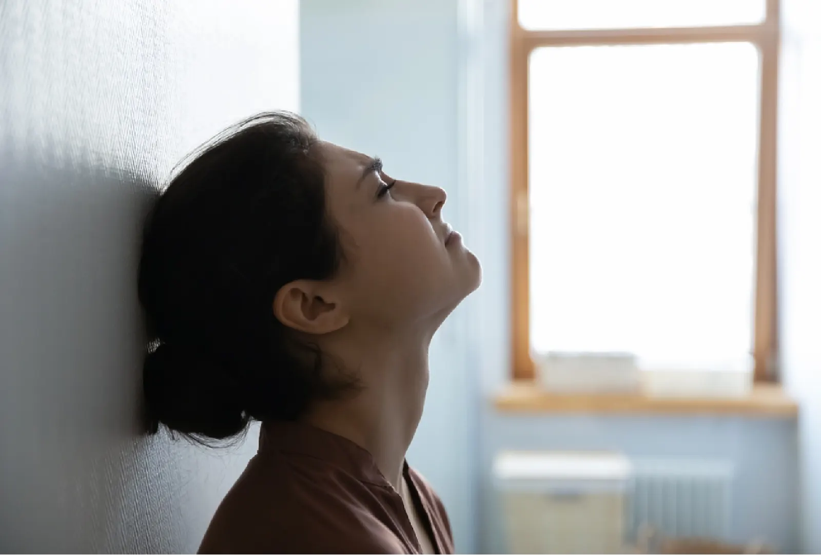 A young woman leans against a wall with her eyes closed, facing the light from a window.